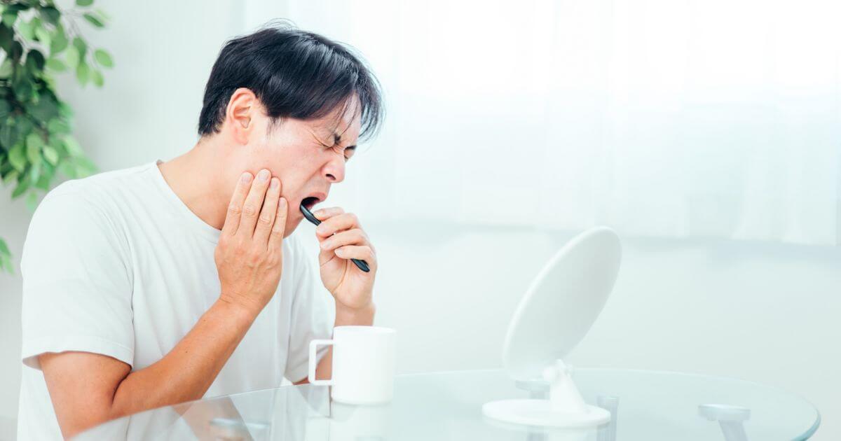 歯の痛みを感じて頬を押さえる男性。歯ブラシを持ち、鏡の前で違和感を確認している。